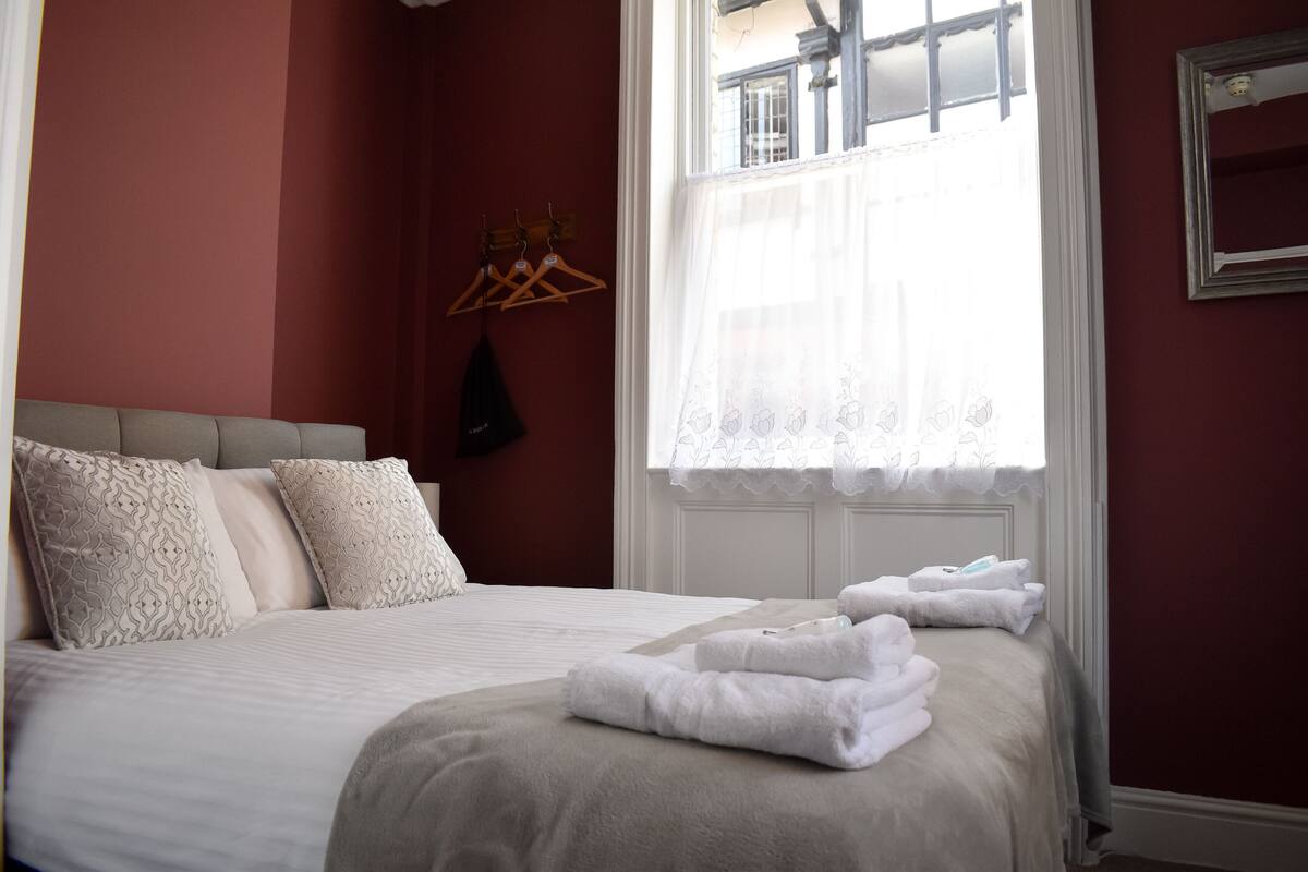 A neatly made bed with white linens and a grey headboard, a window with sheer curtains, and a wall-mounted coat rack with hangers.