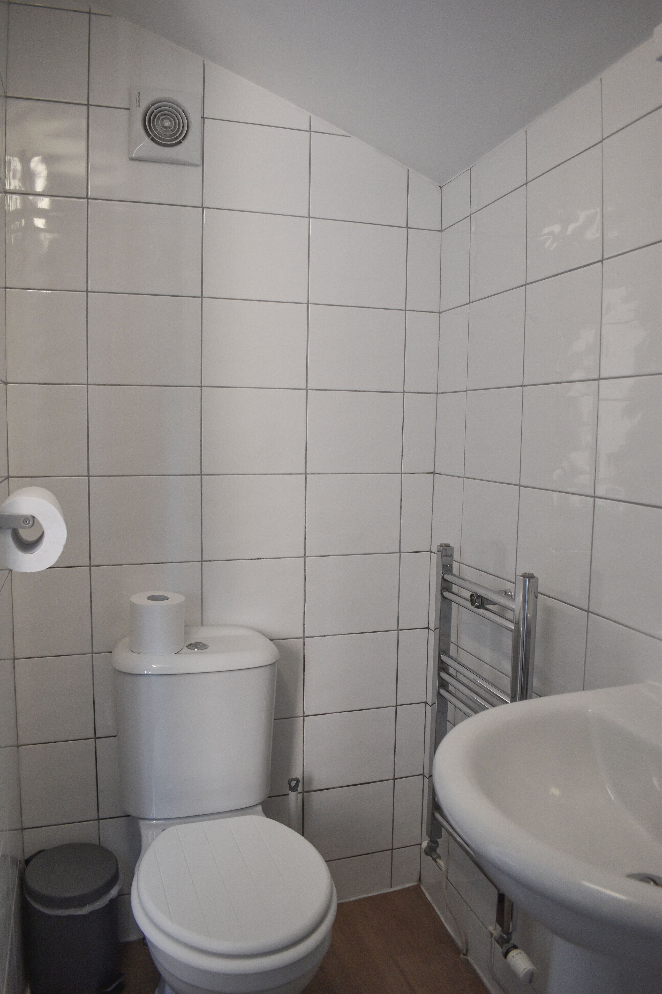 Un baño pequeño y limpio con azulejos blancos, un inodoro, un lavamanos y un cubo de basura.