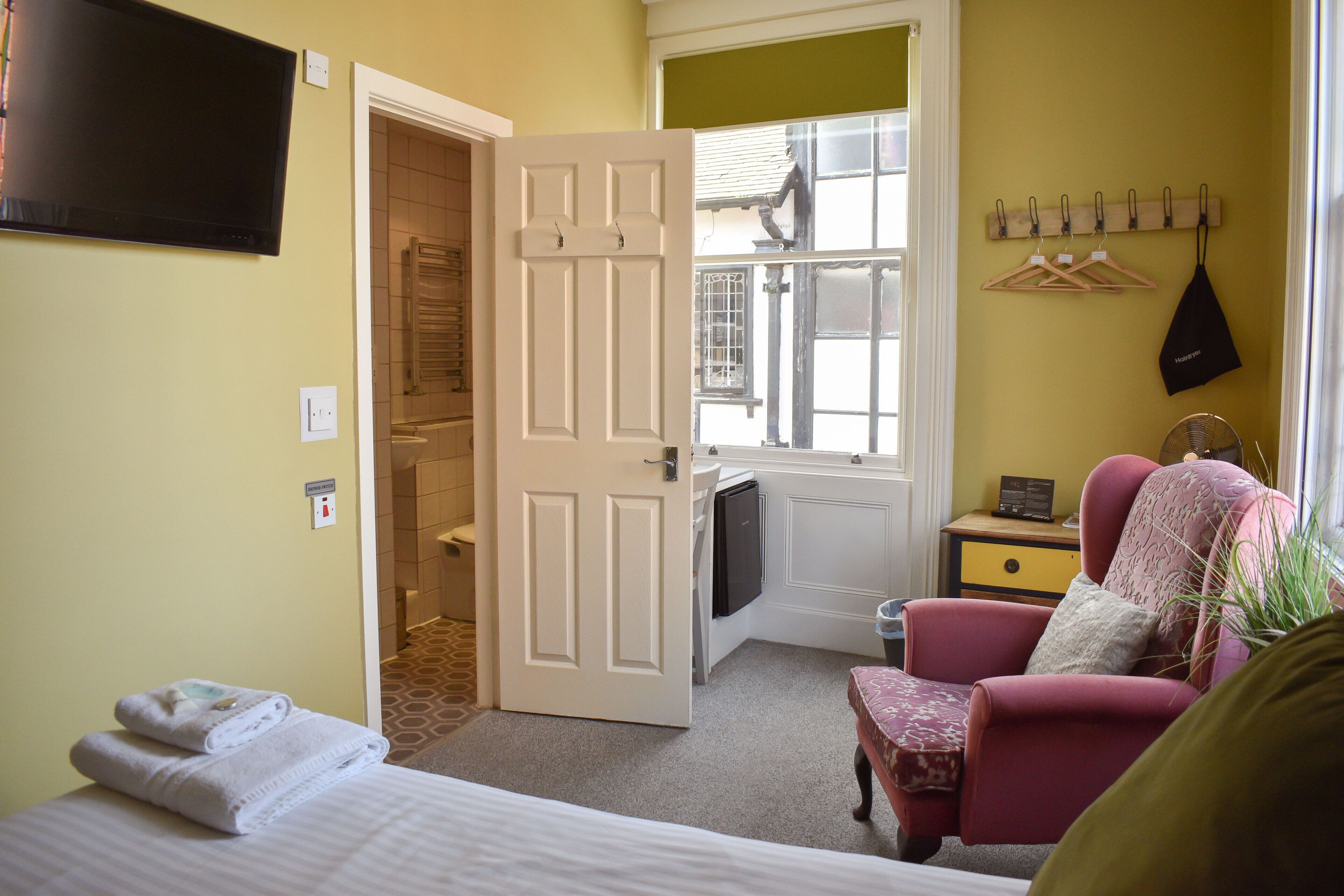 A hotel room with a bed, a chair, a small table, a TV, a window, and a door leading to a bathroom.