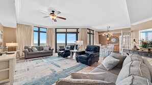 A spacious living room with a large window, a ceiling fan, and a dining area with white furniture. - The Oasis at Orange Beach 1002 (Orange Beach)