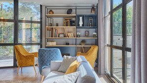 Una sala de estar moderna con un ventanal grande, un sofá, dos sillones y una estantería llena de objetos decorativos.
