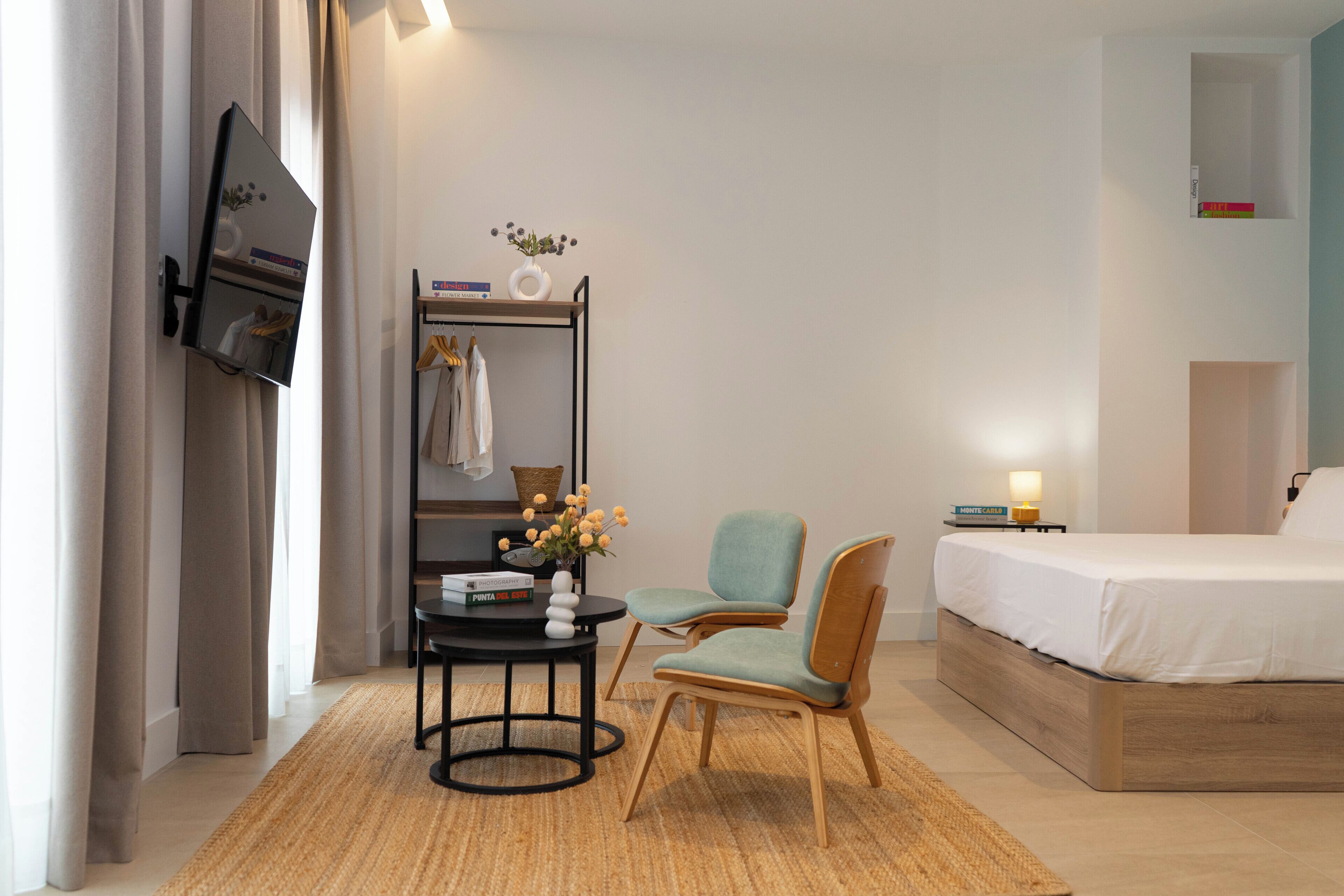 Habitación de hotel con cama, escritorio con silla, una pequeña mesa con un florero y estante con libros y una lámpara.