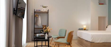 A hotel room with a bed, a desk with a chair, a small table with a vase of flowers, and a shelf with books and a lamp.