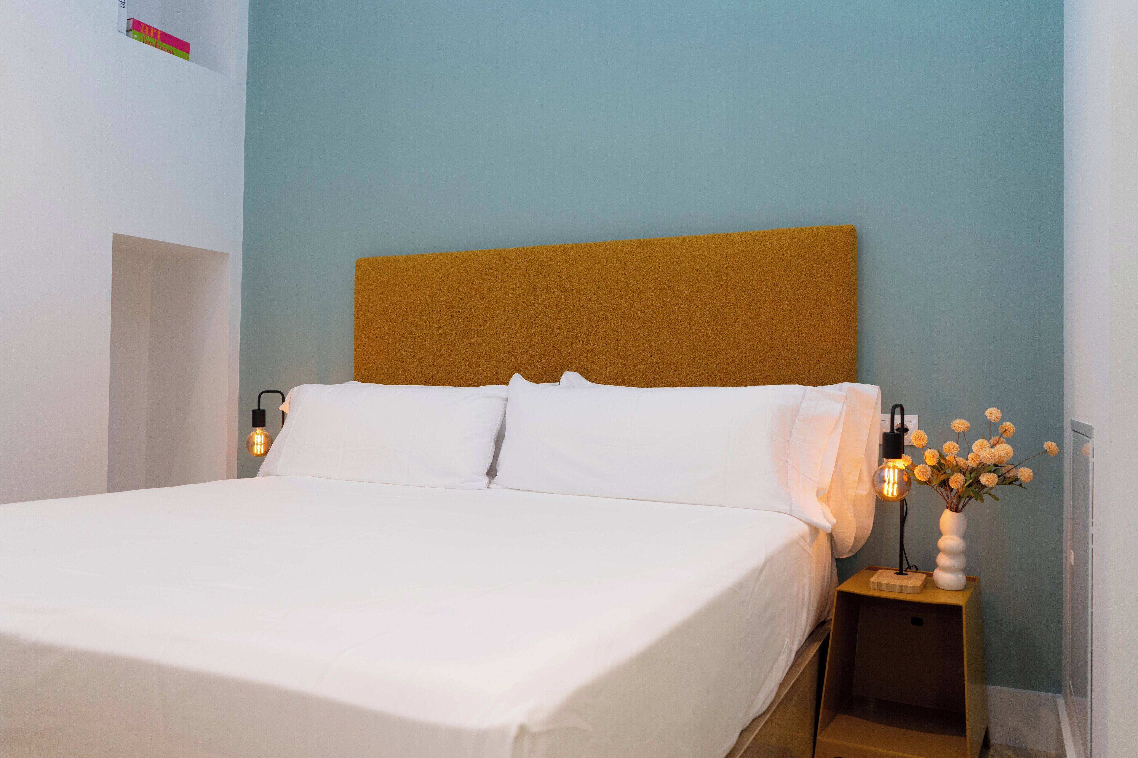 A neatly made bed with white linens, a wooden headboard, and a bedside table with a lamp and a vase of flowers.