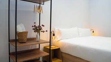 Un dormitorio ordenado con una mesita de noche de madera, un jarrón blanco con flores moradas y una cama con ropa de cama blanca.