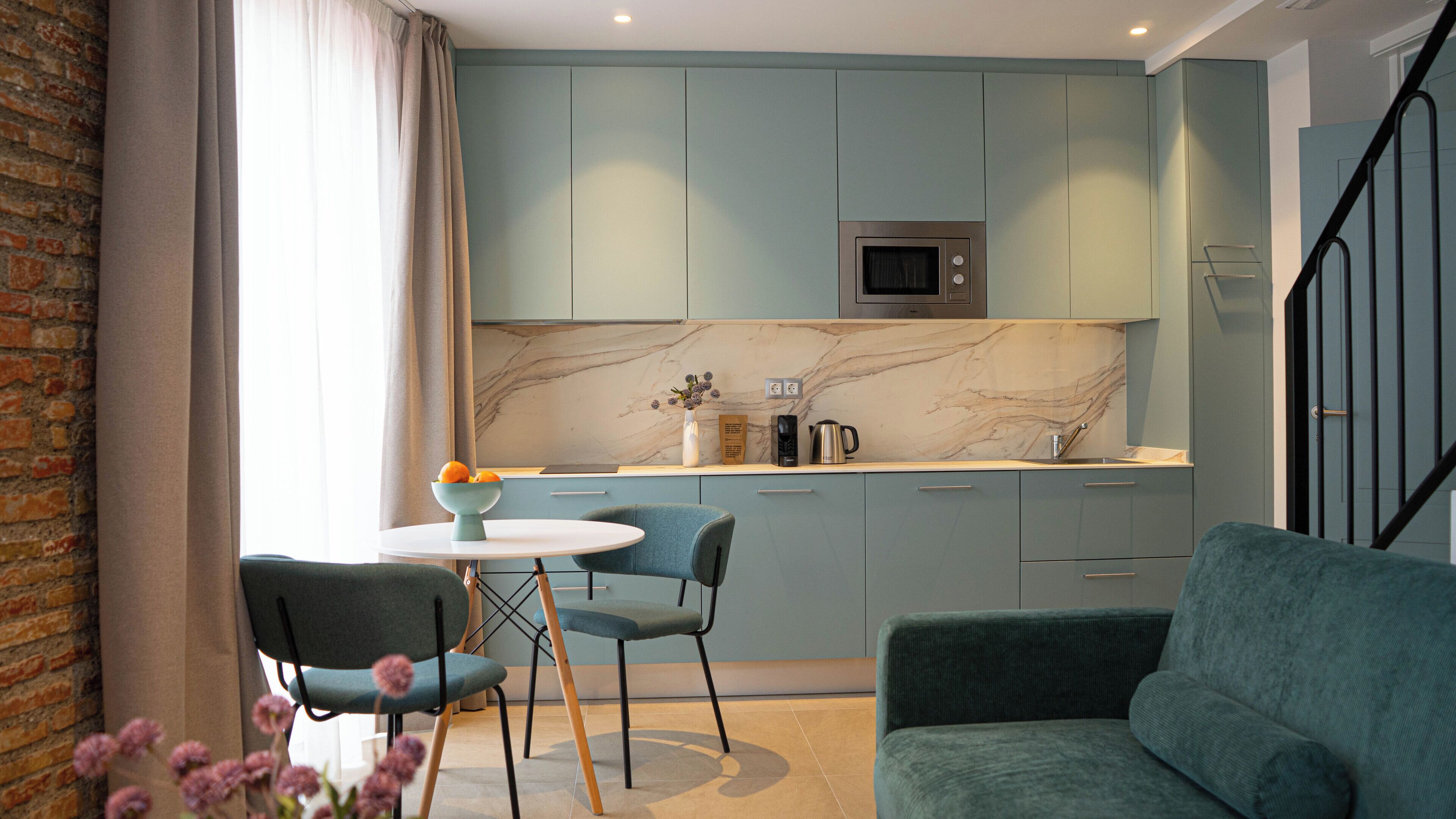 A modern kitchen with teal cabinets, a marble backsplash, a microwave, and a coffee maker. There is a dining area with a round table and two chairs.