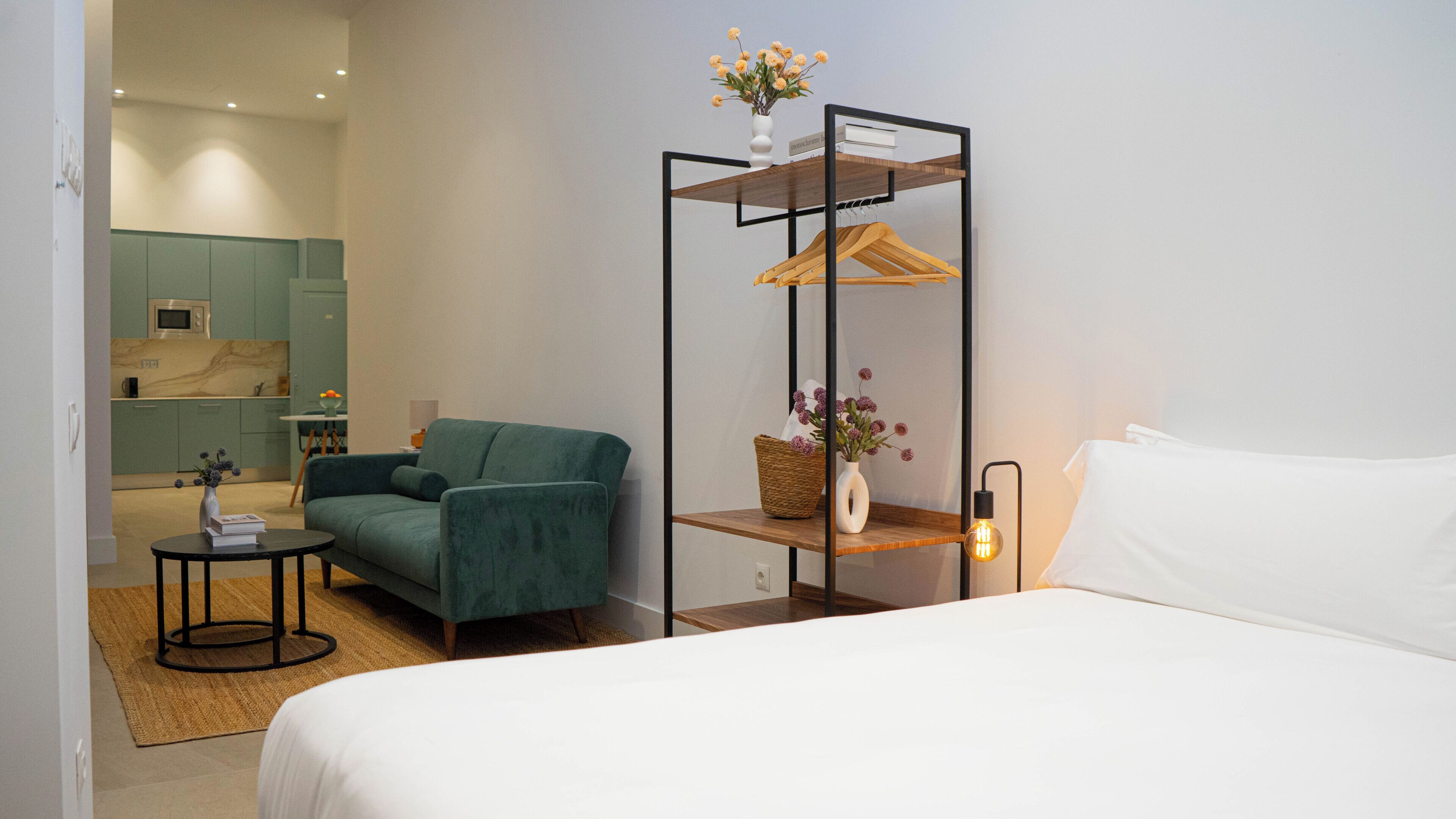 A hotel room with a bed, a green armchair, a small table with a vase, and a wooden shelf with a hanging garment rack.