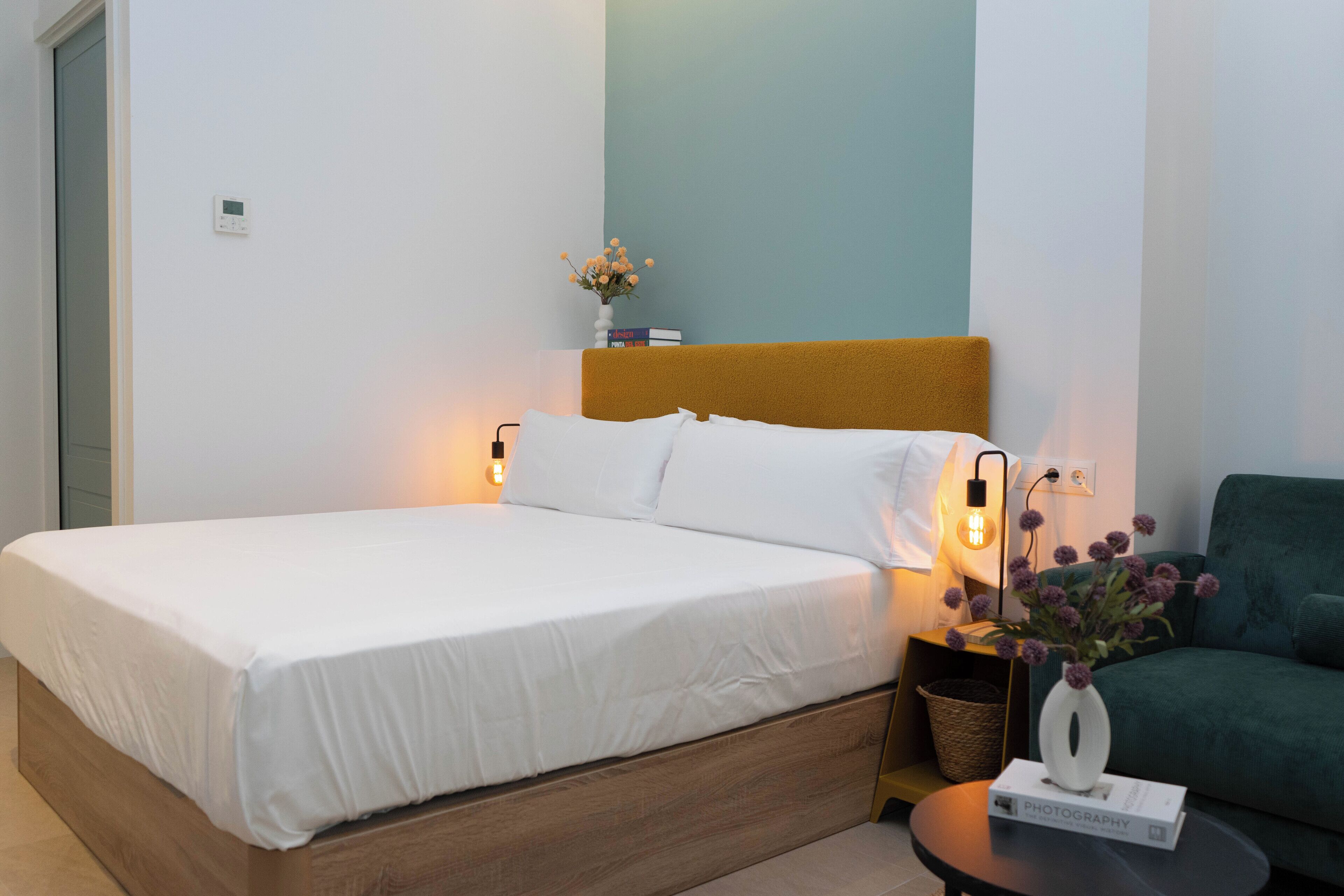 A neatly made bed with white linens, a bedside table with a book and a vase, and a green armchair.