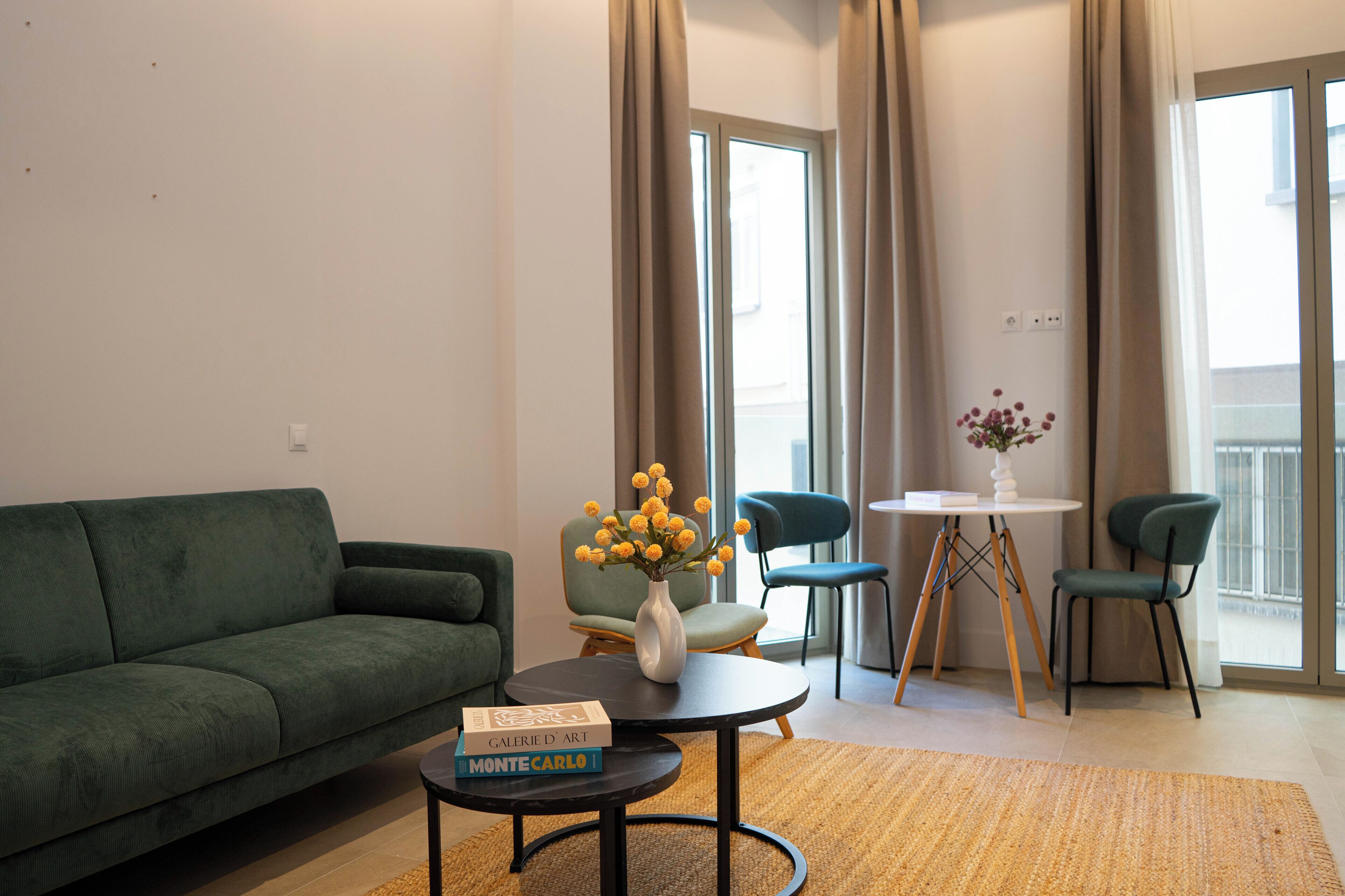 Una sala de estar moderna con un sofá verde, una mesa de centro con libros, un jarrón con flores y un comedor con sillas y una mesa.