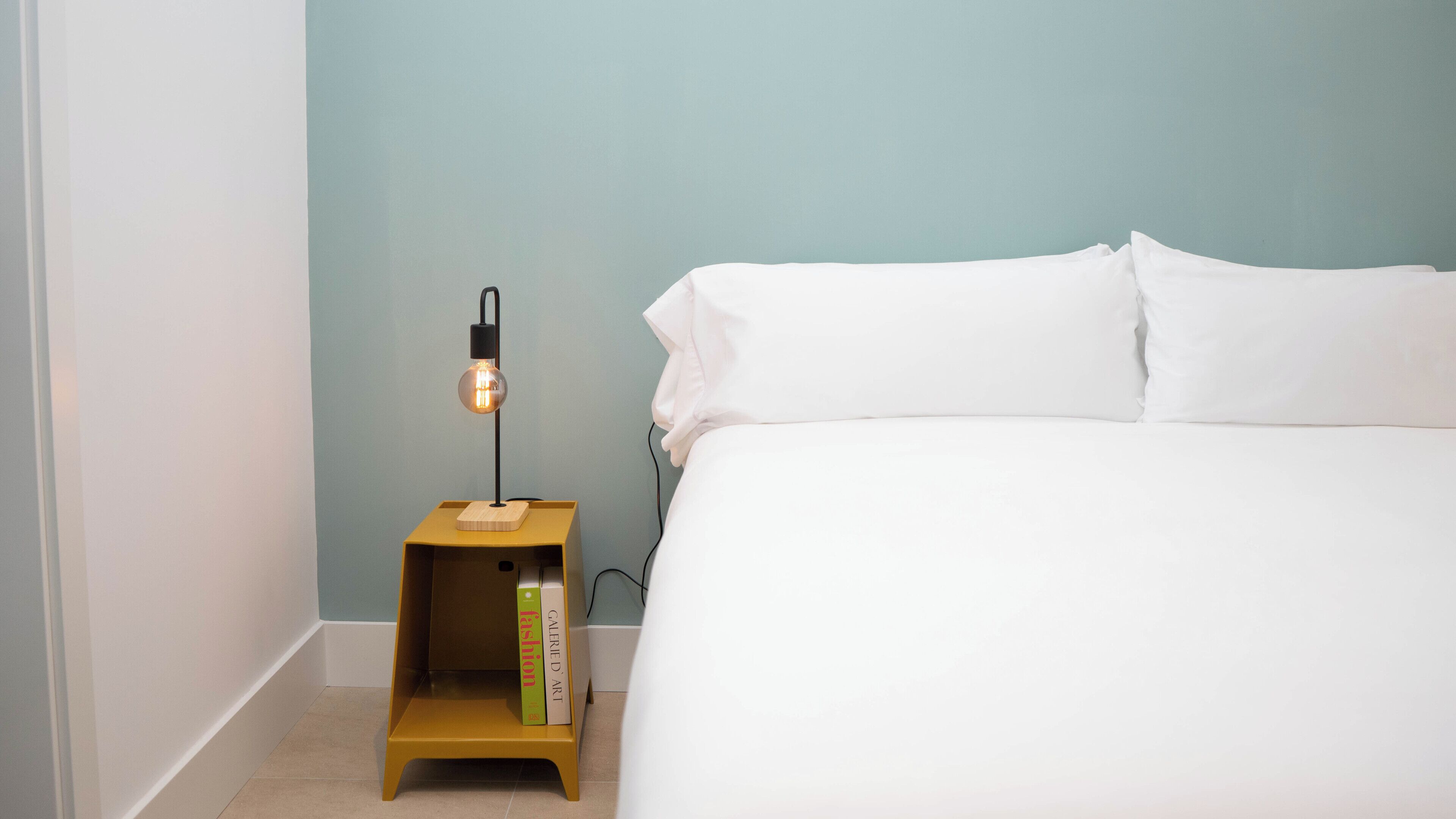 Una cama bien hecha con sábanas blancas, una mesita de noche con lámpara y una pequeña estantería con libros.