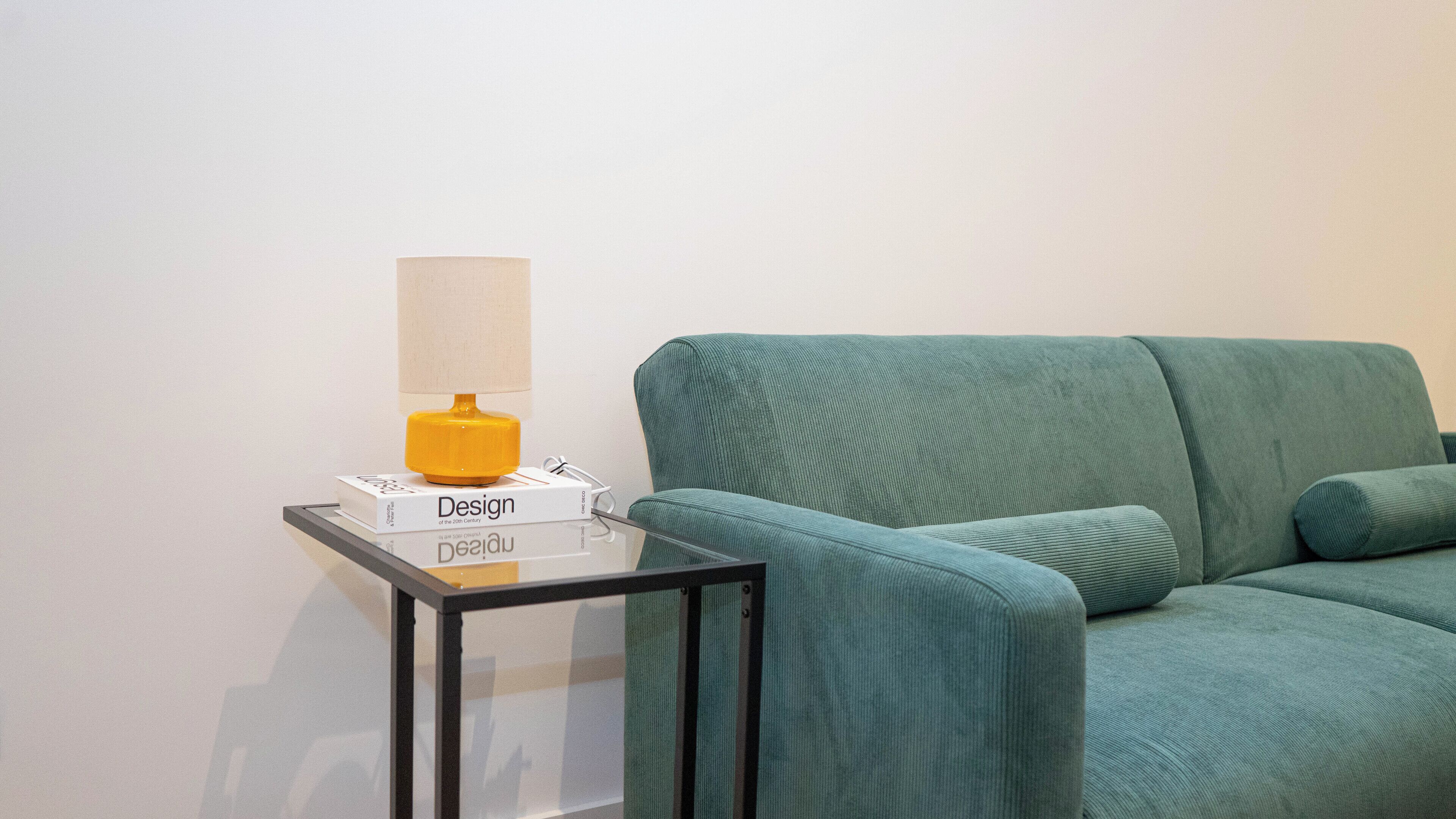 Un fauteuil turquoise est placé à côté d’une table en verre, sur laquelle se trouvent une lampe et un livre intitulé « Le design du XXe siècle ».
