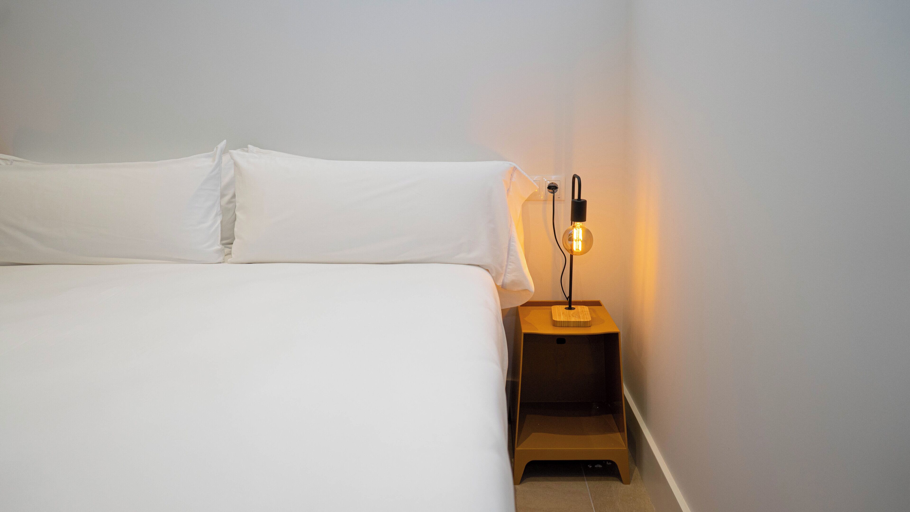 Una cama bien tendida con sábanas blancas en una habitación con una mesita de noche de madera sencilla y una sola bombilla.