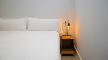 Una cama bien tendida con sábanas blancas en una habitación con una mesita de noche de madera sencilla y una sola bombilla.
