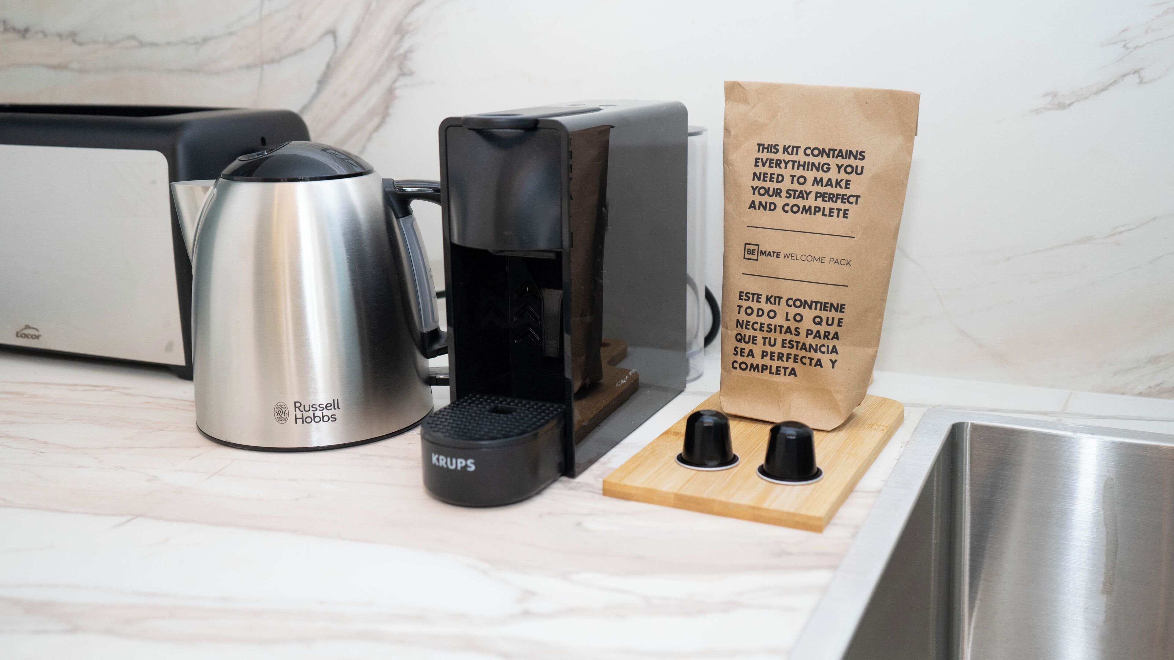 A kitchen with a Russell Hobbs kettle, a Krups coffee maker, and a paper bag labeled "BE MATE WELCOME PACK".