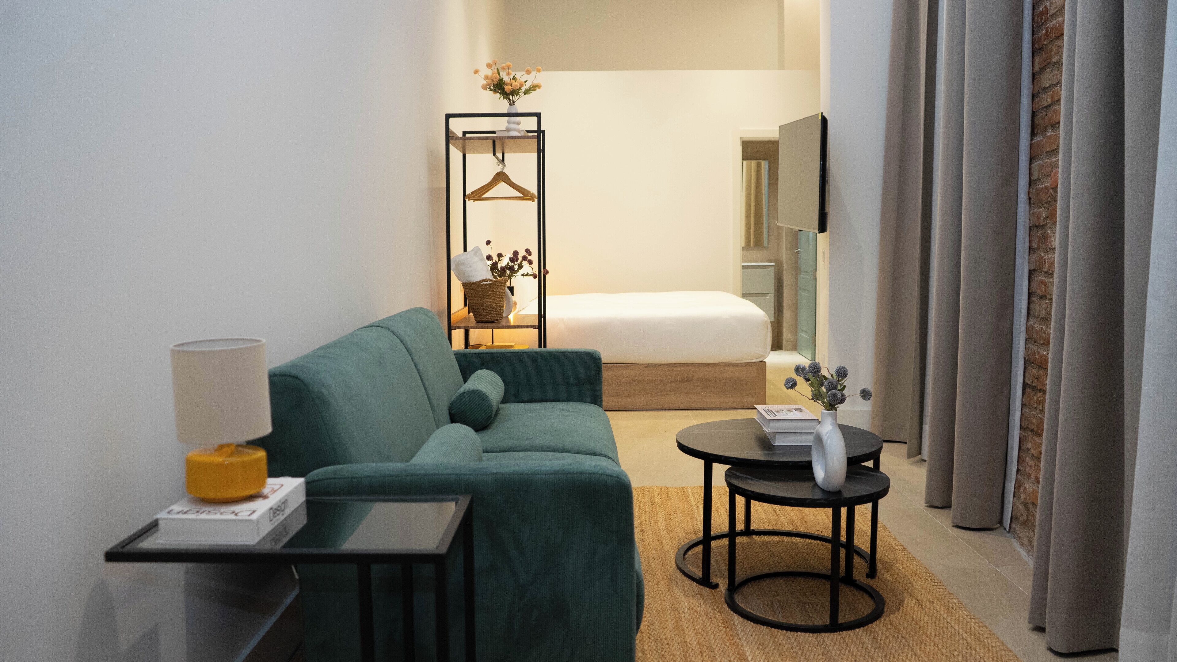 A hotel room with a green sofa, a small table with a lamp, a bedside table with a vase, and a bed with a white comforter.