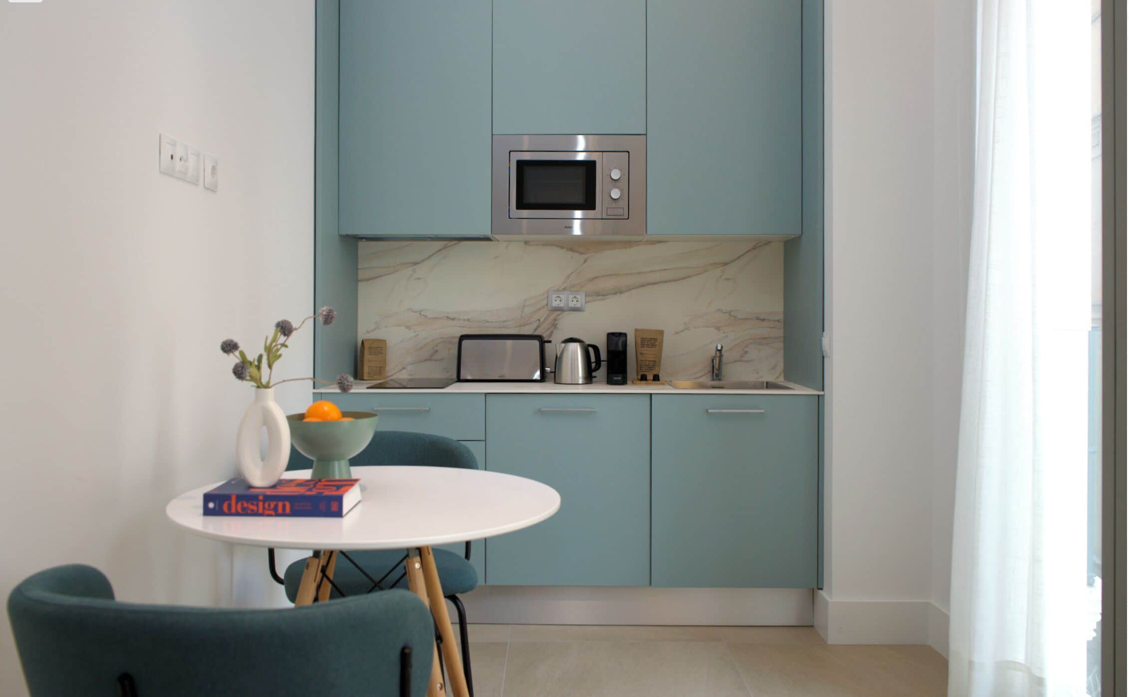 Cocina compacta con armarios color turquesa, encimera blanca, microondas, tostadora y cafetera. Hay una mesa pequeña con un jarrón y un libro.