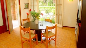 Zona pranzo con tavolo rotondo, quattro sedie di legno, un vaso con fiori e vista all'esterno attraverso un'ampia finestra.