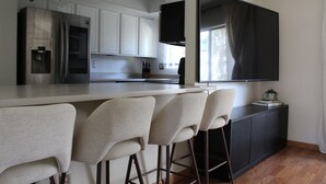 A modern kitchen with white cabinets, a stainless steel refrigerator, a built-in oven, and a large flat-screen TV mounted on the wall.