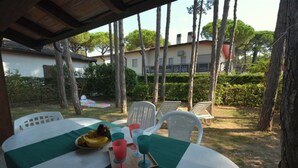 Un área de comedor al aire libre con una mesa preparada para dos, una bandeja con frutas y vasos, y una vista de un edificio y árboles al fondo.