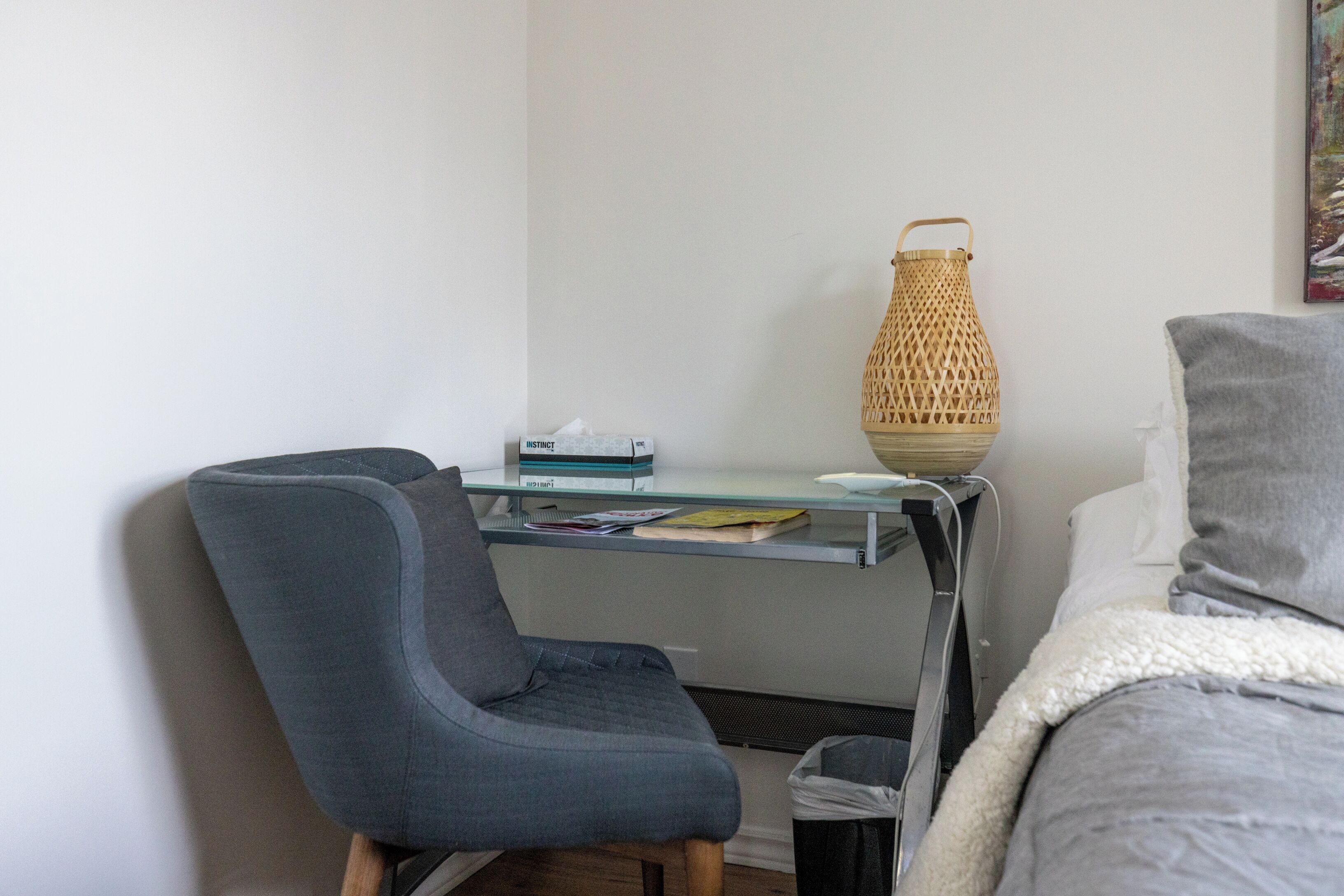 Une pièce avec une chaise grise, un bureau en verre sur lequel sont posés des livres, un panier en osier et un lit recouvert d’une couverture grise.