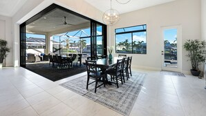 A spacious dining area with a large table and chairs, a ceiling fan, and a view of palm trees through large windows. - Surfside Villa ~ Entire home in Cape Coral, Florida (Cape Coral)