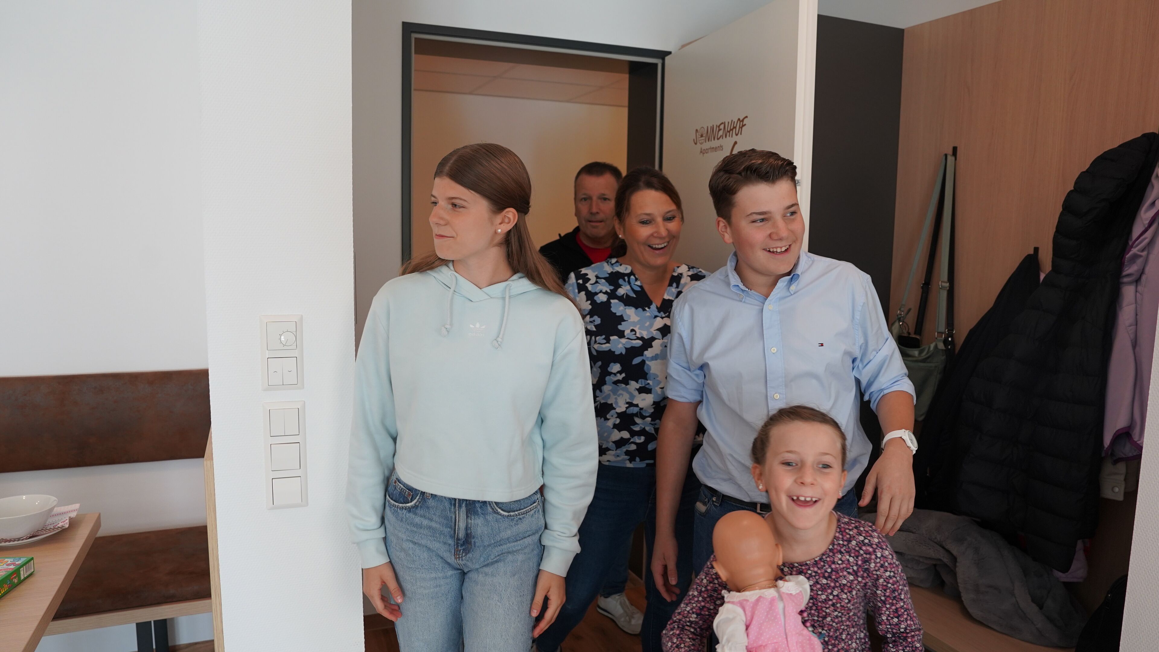 Eine vierköpfige Familie in einem modernen Wohnzimmer mit einer Holzbank, einem Esstisch mit Schüssel und einer Wand mit einem Schild.