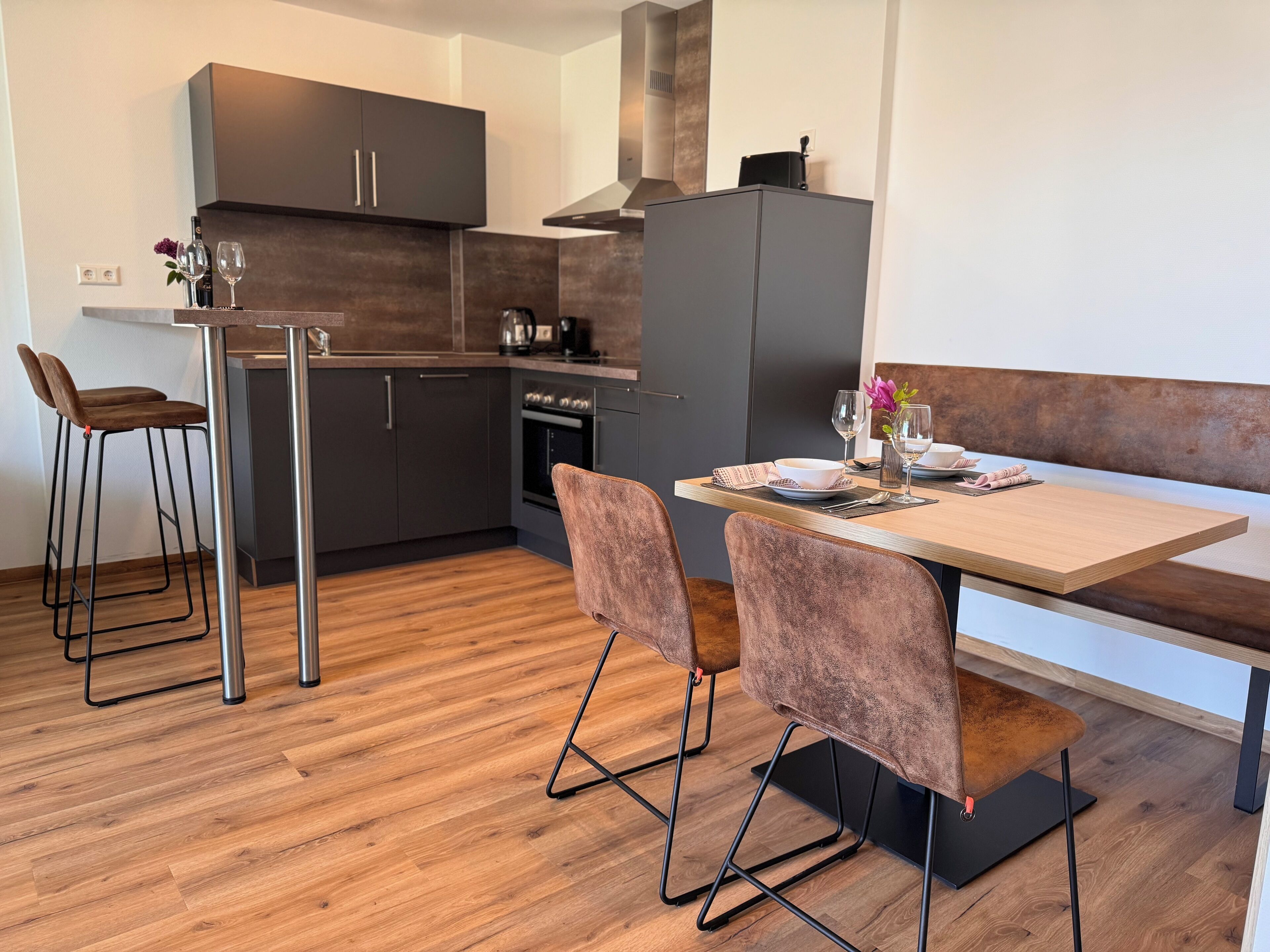 Une cuisine moderne avec des meubles de couleur sombre, un îlot central doté de tabourets et une table à manger pour deux personnes.