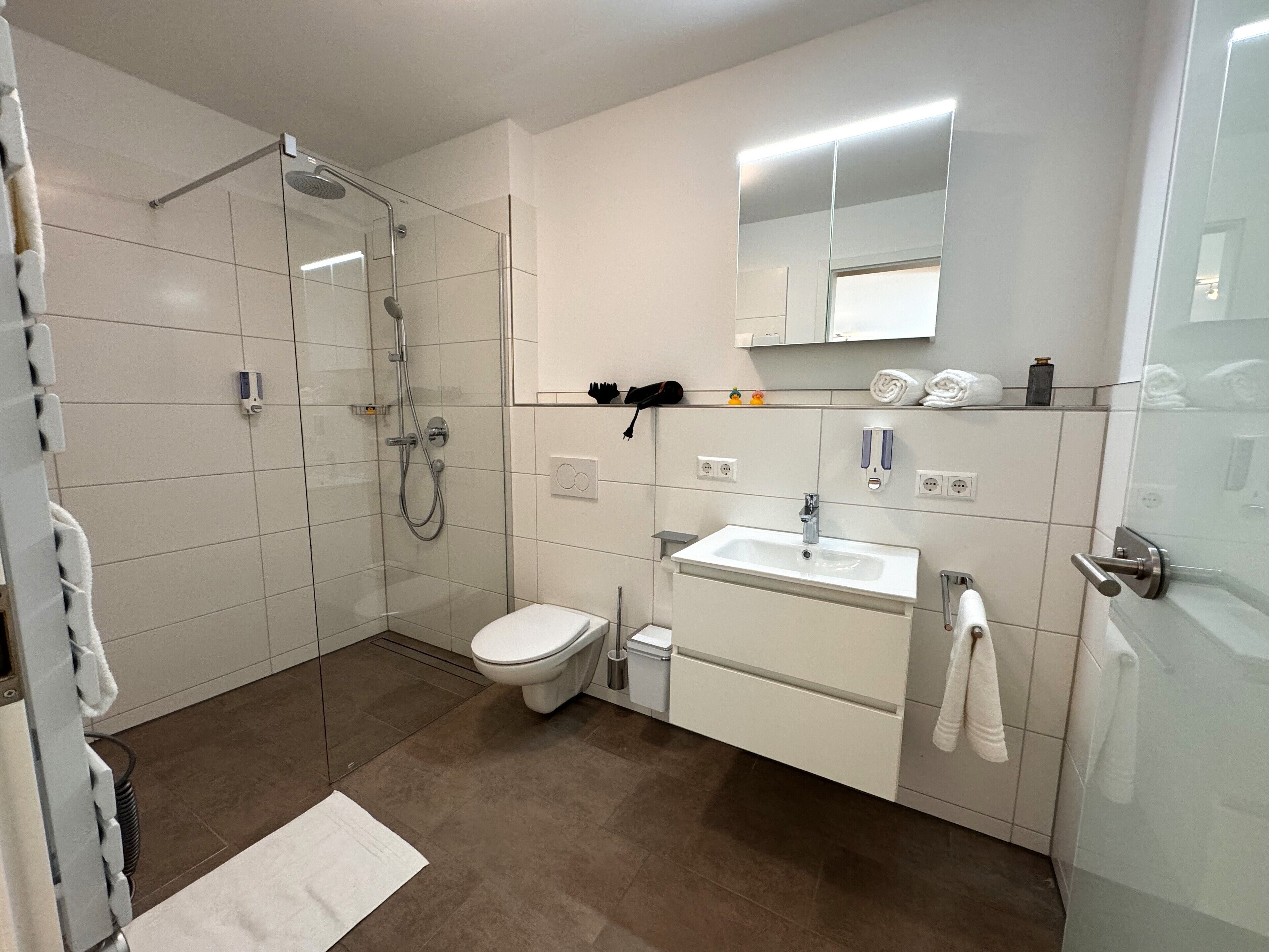 Un baño moderno con cabina de ducha de vidrio, inodoro blanco, lavamanos con espejo y un perchero para toallas.