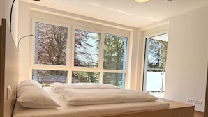 A modern bedroom with a large window, a bed with white linens, and a bedside table with a book. - SONNENHOF Apartments (Bad Duerrheim)