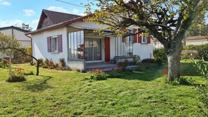 A single-story house with a front porch, a small garden with red flowers, and a tree in the foreground. - Quiet, flower-filled house in the countryside, good location, easy access (Le Grand-Lemps)