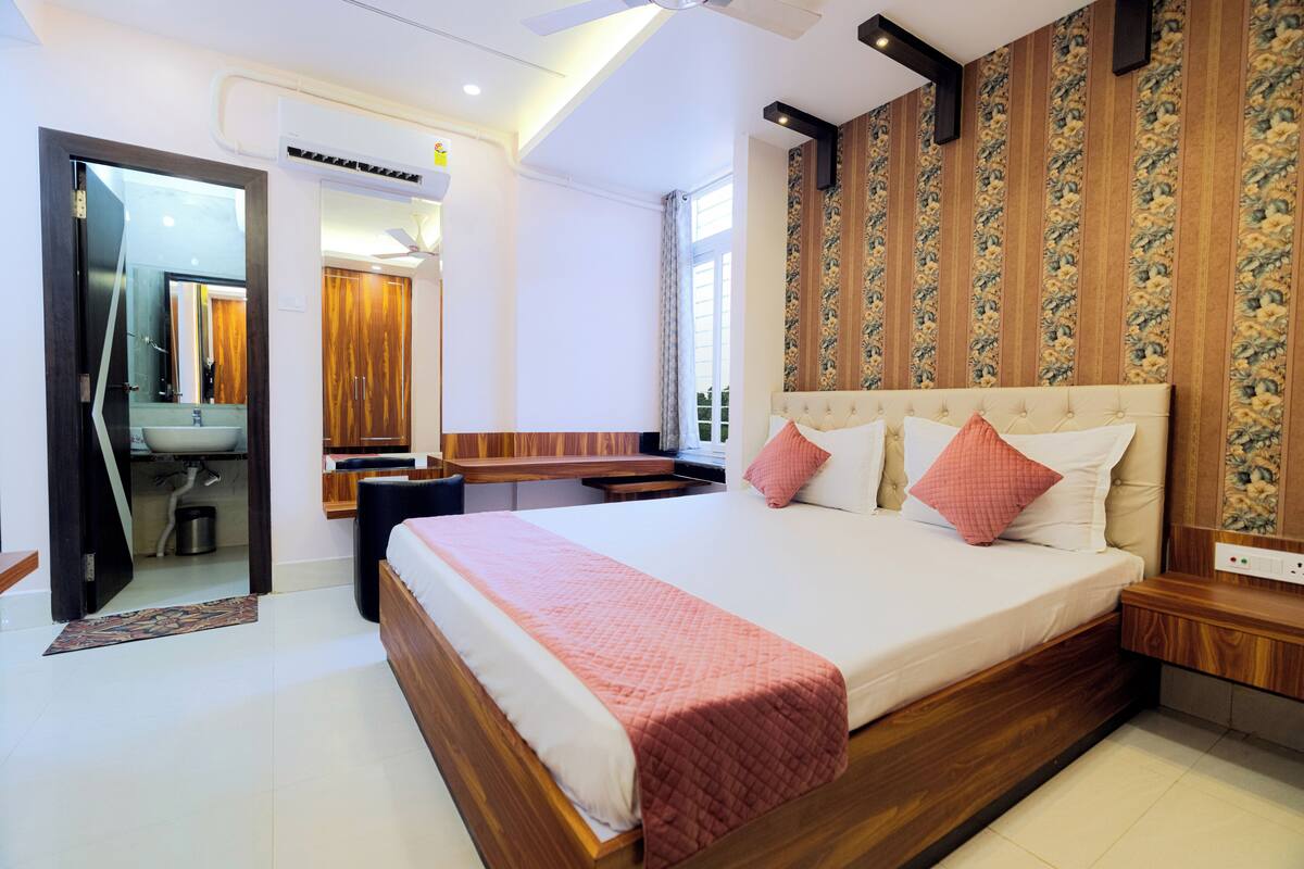 A modern bedroom with a large bed, a wooden headboard, a bedside table, a nightstand, a mirror, a vanity, and a window with curtains.