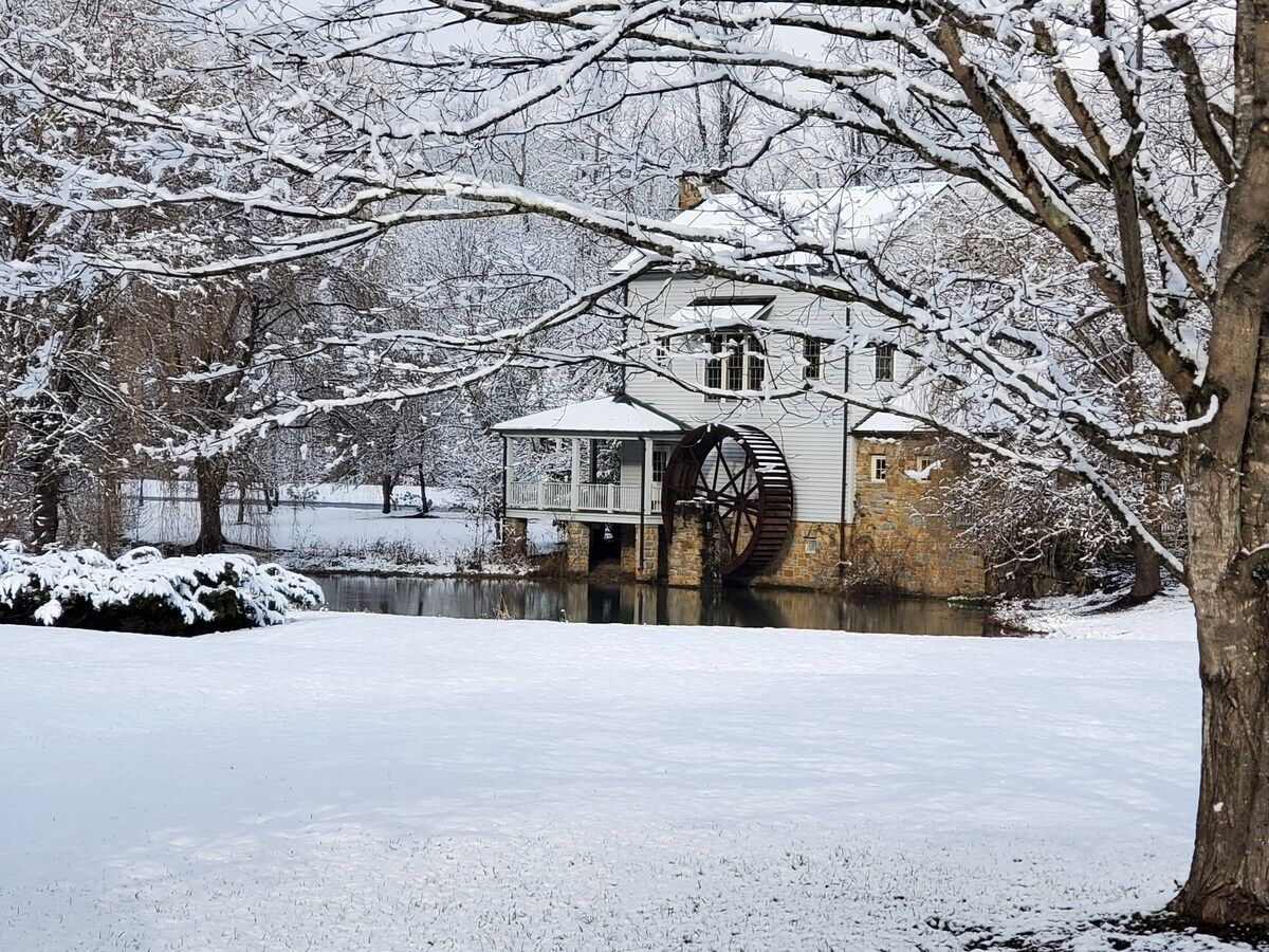A historic building with a large water wheel, surrounded by snow-covered trees and a frozen pond.