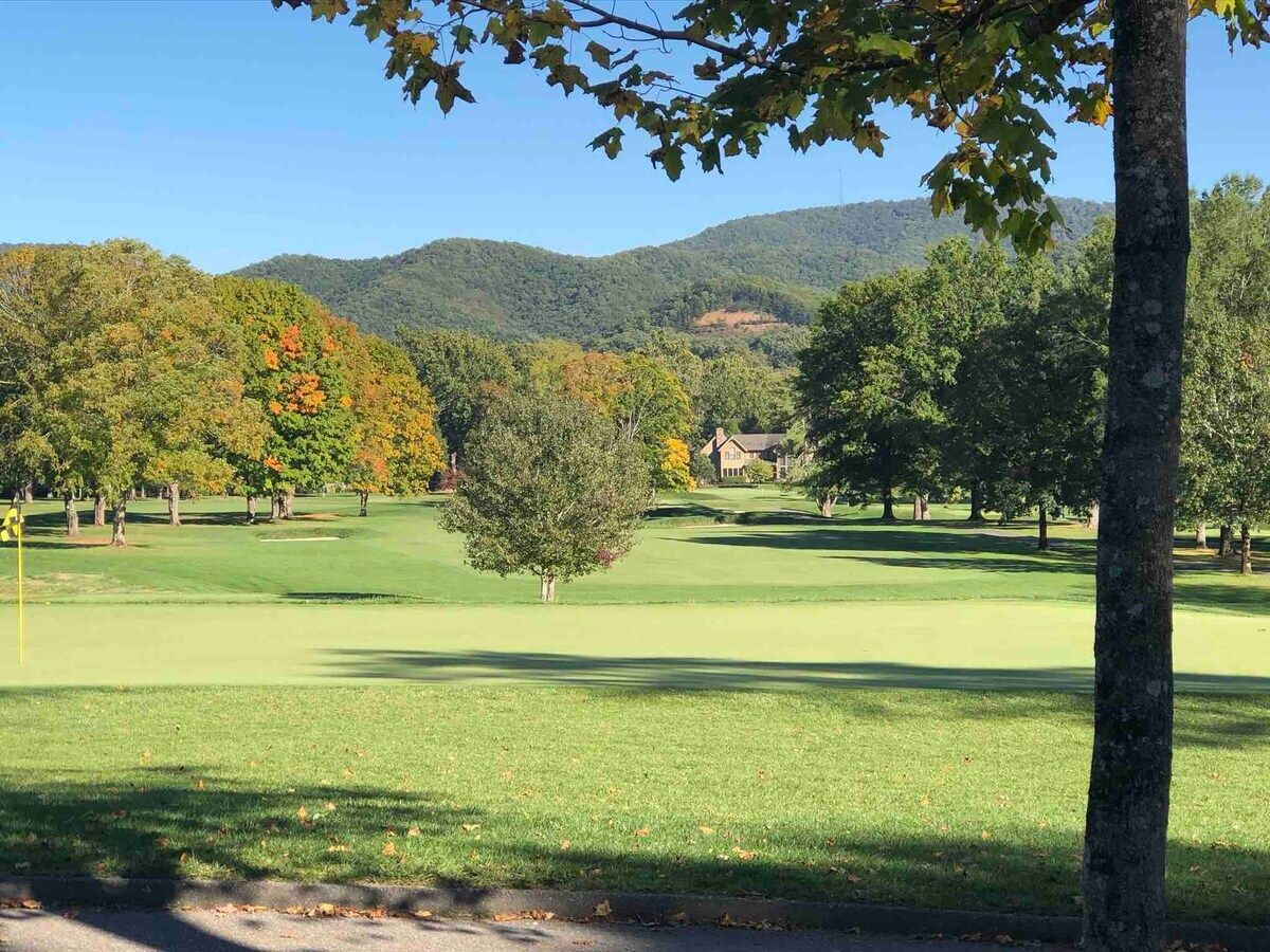 A golf course with a clear sky, trees with autumn foliage, and a distant house.