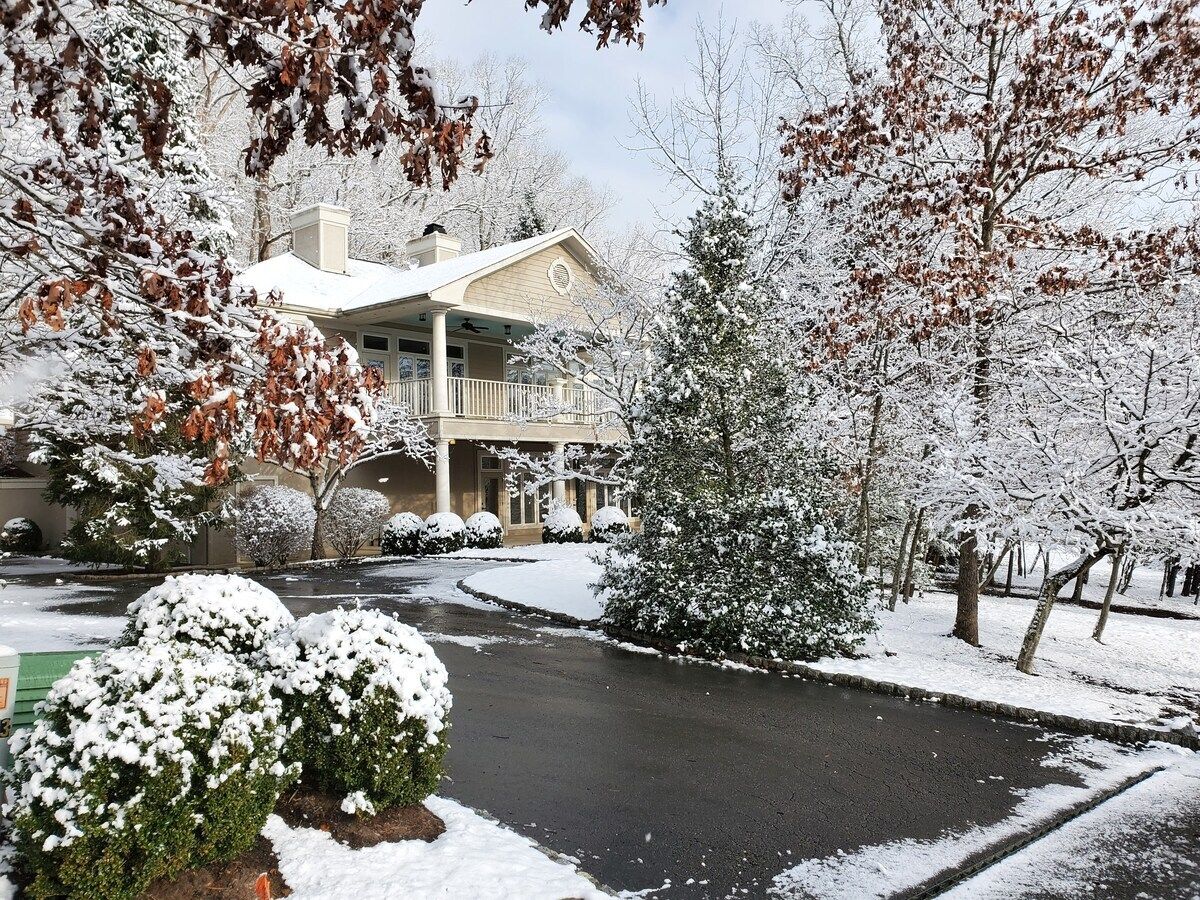 A residential property with a covered driveway, snow-covered trees, and a two-story house with a balcony.