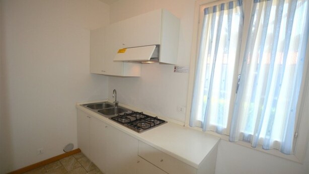 A small kitchen with white cabinets, a gas stove, and a window with sheer curtains. - Charming Villa Near the Beach With Parking (Lignano Sabbiadoro)