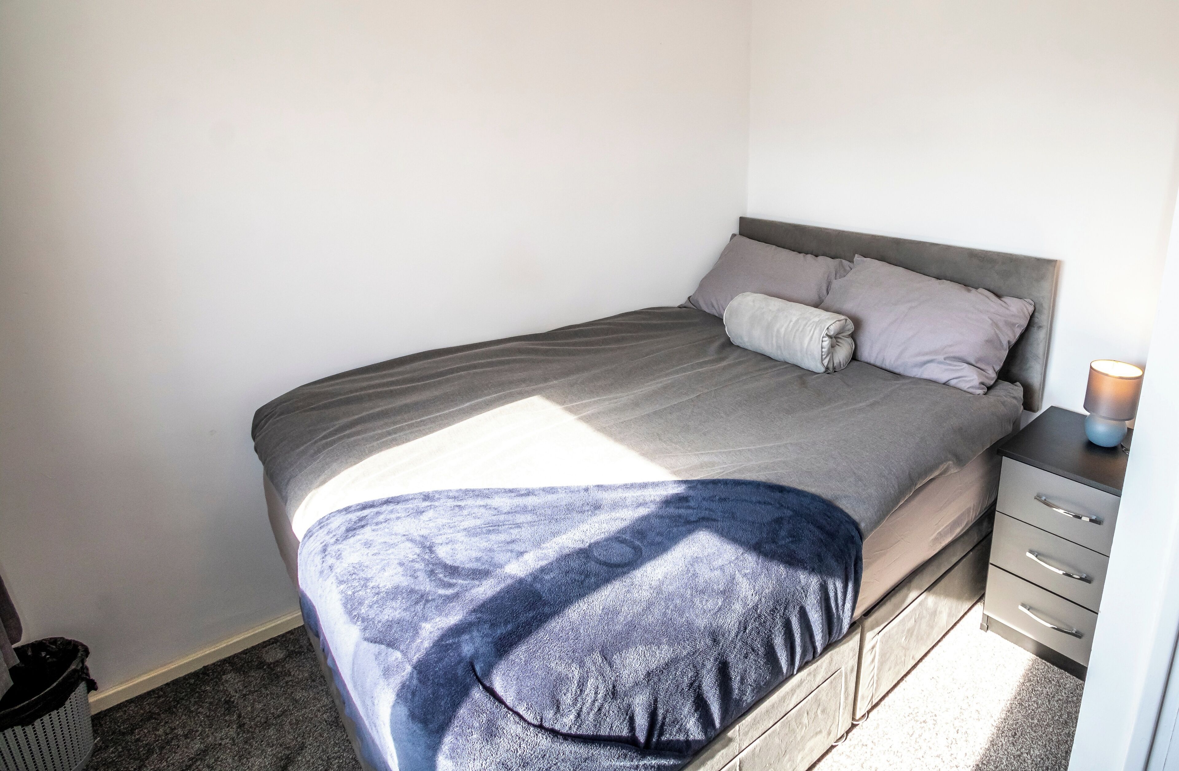 Een net opgemaakt bed met een grijze hoofdbord en een nachtkastje met een lamp.