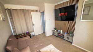 A living room with a brown sofa, a TV mounted on a wall, and a door leading to another room. - 3 Bed Home - Sleeps 8 - Stoke City fc - Parking (Stoke-on-Trent)