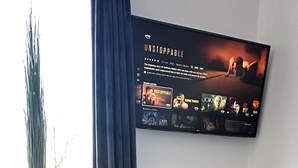 A modern living room with a white table, a remote control, and a candle on a white rug. A flat-screen TV mounted on the wall displays a movie poster. - HARMONY "4" 1-room apartment with kitchen (Gütersloh)