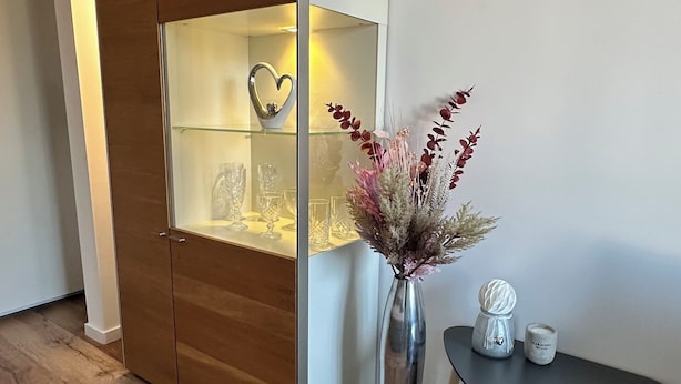 A room with a wooden wardrobe, a glass display cabinet, a vase with dried flowers, and a small round table with a candle. - HARMONY "4" 1-room apartment with kitchen (Gütersloh)