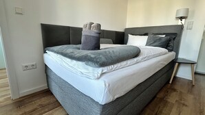 A neatly made bed with a grey and white duvet, a grey throw blanket, and a pair of grey slippers on the footboard. - HARMONY "4" 1-room apartment with kitchen (Gütersloh)