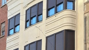 A multi-story building with a beige facade, large windows with dark shutters, and a ground-level entrance with a wooden door. - Apartamentos Turísticos en Albacete (Albacete)