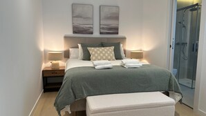 A modern bedroom with a bed, bedside tables, a bench, and a glass-enclosed shower. - Apartamentos Turísticos en Albacete (Albacete)