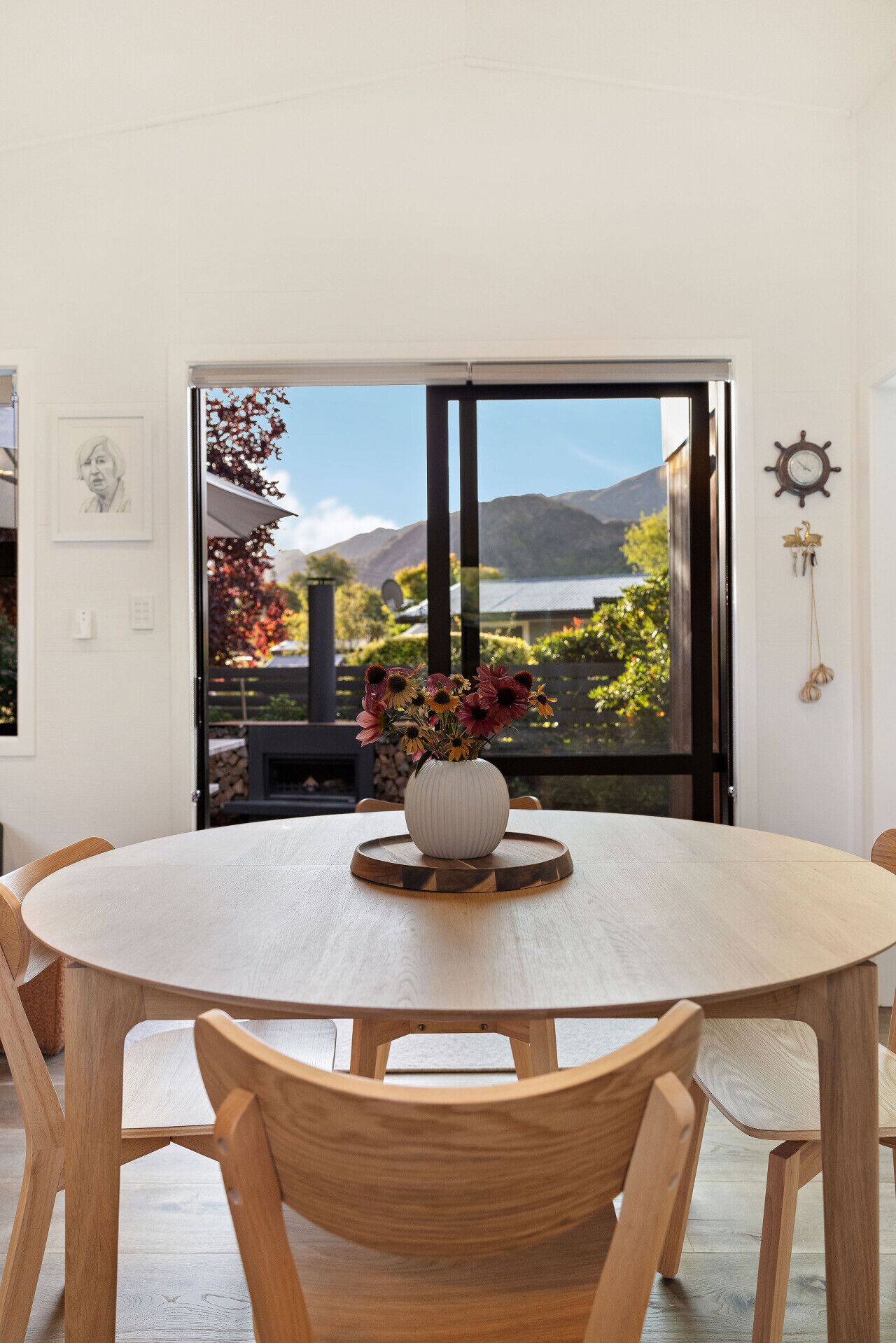 Un tavolo rotondo in legno con un vaso bianco pieno di fiori, con vista su un paesaggio suggestivo attraverso una grande finestra.