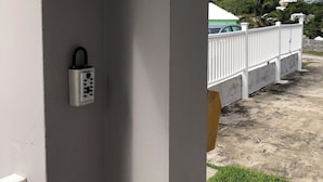 A view from a balcony with a white railing, a security keypad, and a glimpse of a green car and houses in the background. - Charming Central 1-Bedroom Apartment Close To City Of Hamilton (Devonshire Parish)