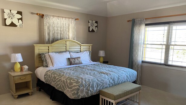A bedroom with a bed, bedside tables, a bench, a window with curtains, and wall art. - Charming Central 1-Bedroom Apartment Close To City Of Hamilton (Devonshire Parish)