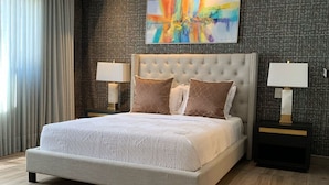 A modern bedroom with a tufted headboard, a large bed with patterned bedding, two bedside tables with lamps, and a colorful abstract painting on the wall. - Breakfast Included Regatta Living II 404 (Santo Domingo)