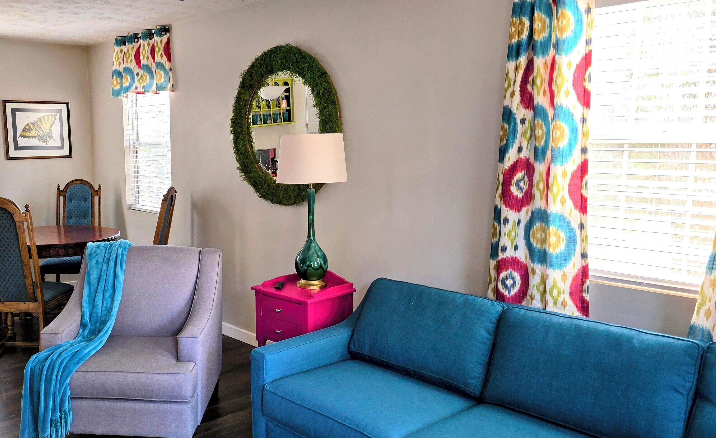 A living room with a blue sofa, a grey armchair with a blue throw, a pink side table, a green lamp, and a wall-mounted mirror with a green wreath.