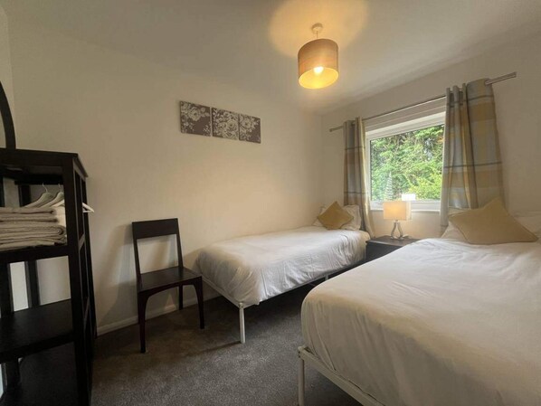 Un dormitorio con dos camas individuales, una silla, una mesita de noche con lámpara y una ventana con cortinas.
