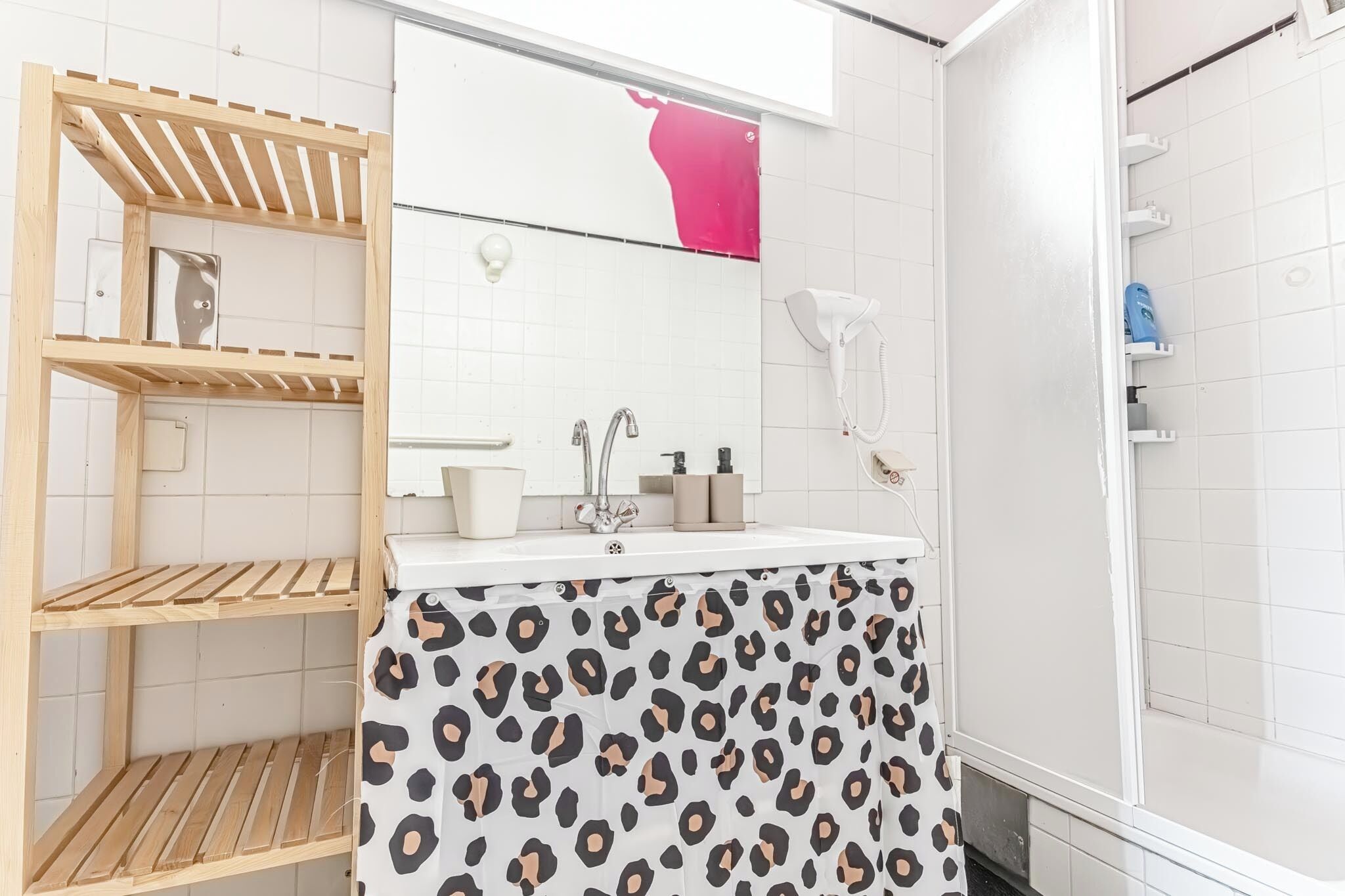 A bathroom with a wooden shelf unit, a sink with a mirror, a shower area, and a leopard print shower curtain.