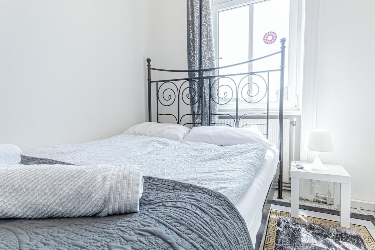 A bed with a metal frame, white bedding, and a small bedside table with a lamp.